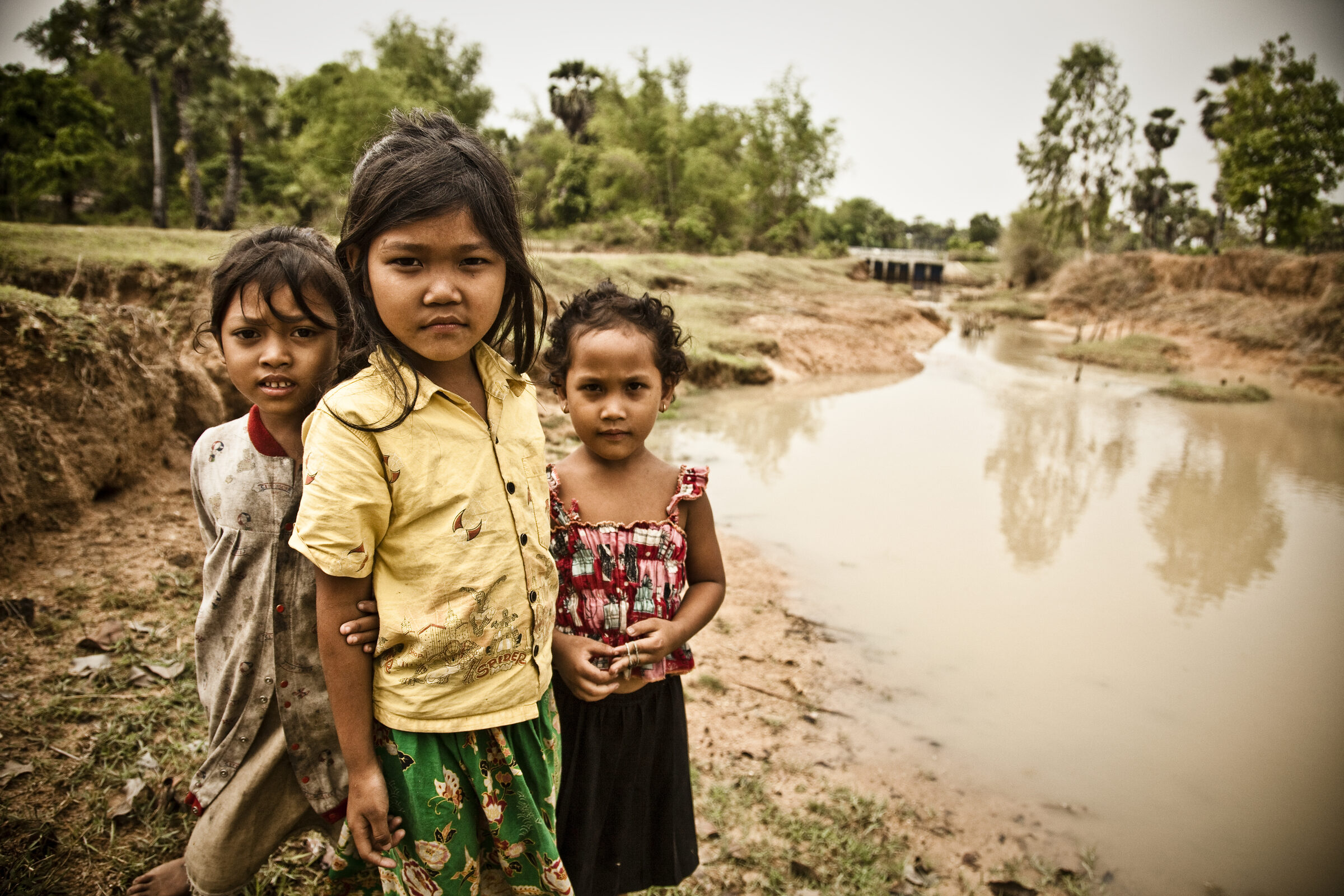 Three children standing beside a low river at the end of dry season