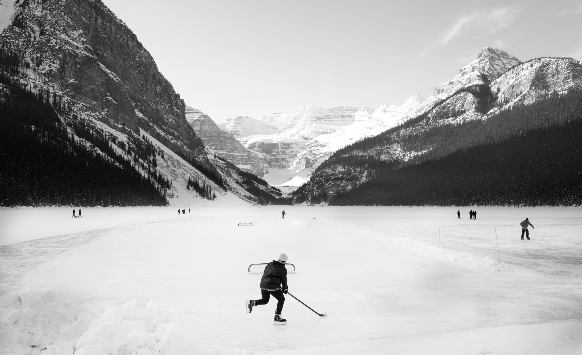 A lone hockey player skating on a cleared patch of a frozen lake surrounded by towering snow-dusted mountains